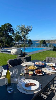 an outdoor dining table with food and wine on it, overlooking the water in front of a house