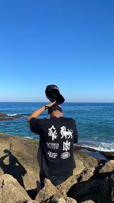 a man standing on top of a rock next to the ocean with a hat on his head