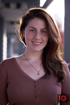 a woman with long hair smiling at the camera and wearing a brown shirt, standing in front of a building