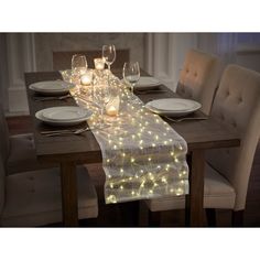 a dining table set with white plates and glasses on it, lit up by fairy lights