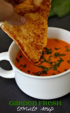 a hand dipping a piece of bread into a bowl of tomato soup with fresh herbs