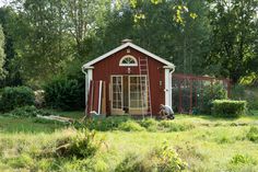 a small red house in the middle of a field with trees and bushes around it