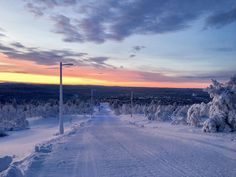 a snow covered road with trees on both sides and sunset in the distance behind it