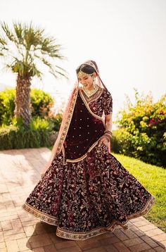 a woman in a maroon and gold bridal gown standing on a brick walkway with palm trees