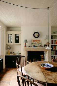 a dining room table and chairs with a clock on the wall above it in front of a fireplace