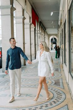 an engaged couple holding hands and walking down the sidewalk in front of a building on a sunny day