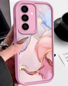 a woman holding up her phone case with pink and gold marble design on it, while sitting