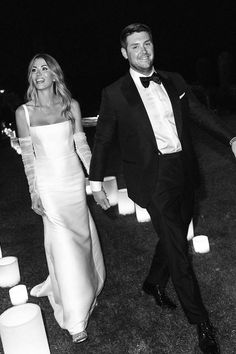 a man and woman in formal wear walking through the grass at night with candles behind them