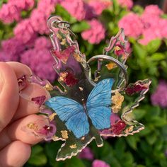 a hand holding a blue butterfly shaped brooch in front of pink and purple flowers