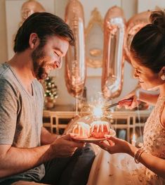 a man and woman sitting on a bed looking at a cake with candles in it