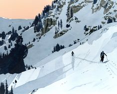 two skiers skiing down a snowy mountain slope at sunset with trees in the foreground