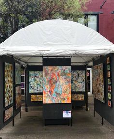 several paintings are displayed under a white tent on the sidewalk in front of a building