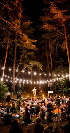 an outdoor concert in the woods with string lights strung over it and people sitting on the ground