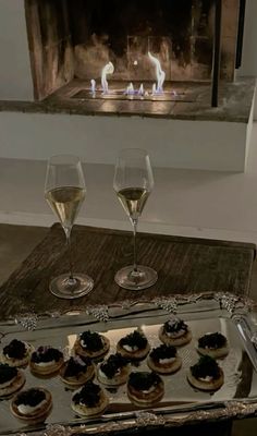 two glasses of wine and some food on a tray in front of a fire place