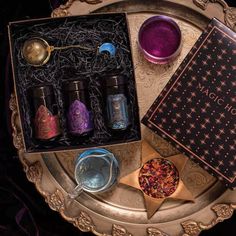 A decorative tray holds a gift box labeled Gemstone Traveler Gift Set by Magic Hour containing three black bottles with colorful labels, a tea infuser in the shape of a sphere, and a crescent moon charm. Surrounding the box are a purple candle, blue liquid in a cup, and a star-shaped dish with dried petals. Healthy Gift Basket, Sampler Box, Tea Sampler, Magic Hour, Natural Wine, Random Ideas, Jar Gifts, Pretty Stuff, Spiritual Jewelry