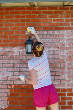 a woman in pink shorts painting a brick wall with a yellow sprayer on it