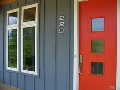 a red door is next to a gray house with two windows and a number on it