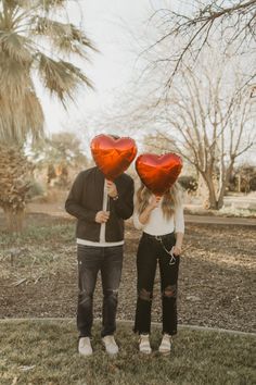 two people standing in the grass holding red heart shaped balloons over their heads with trees behind them