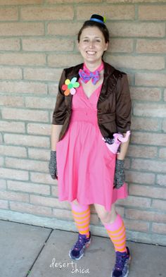 a woman in a pink dress and brown jacket standing next to a brick wall with her hands on her hips