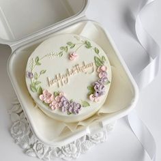 a birthday cake in a white box with pink and purple flowers on the side, sitting on a lace doily