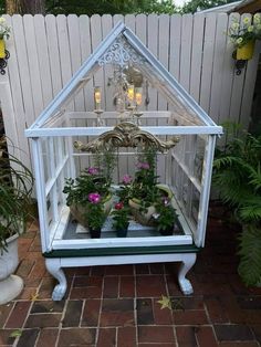 a white greenhouse with potted plants in it on a brick patio next to a fence