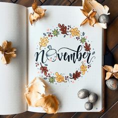 an open book with the word november written in cursive writing on it, surrounded by autumn leaves and acorns