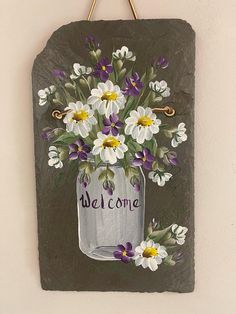 a painted mason jar with white and purple flowers on it that says welcome hanging on a wall