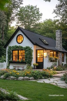 a small white house with windows and plants on the front porch, surrounded by greenery