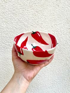 a hand holding a red and white bowl with peppers painted on the bottom, in front of a white wall