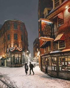 two people walking down a snowy street in the middle of an old town at night