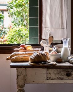 an old table with bread, milk and other items on it in front of a window