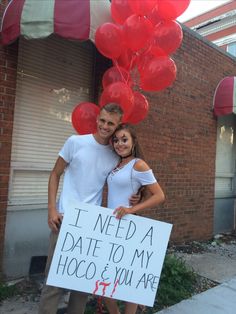 a man and woman standing next to each other in front of a building holding a sign that says i need a date to my hoco & you are it