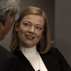 a smiling woman in a black suit and white shirt is talking to an older man