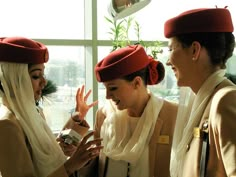 three women wearing red hats and scarves are looking at their cell phones while standing in front of a window