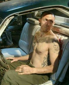 a shirtless man sitting in the driver's seat of a car with tattoos on his chest