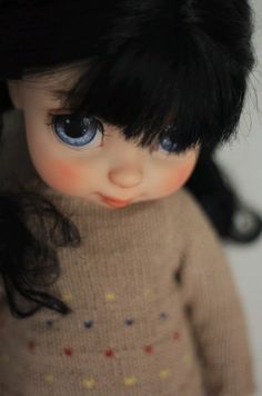 a doll with black hair and blue eyes wearing a brown sweater is standing in front of a white wall