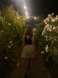 a woman walking down a path next to lots of flowers at night with lights on