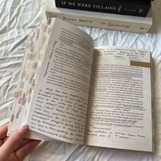 a hand holding an open book on top of a bed next to two stacks of books