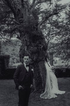 a man standing next to a woman in front of a large tree with vines on it