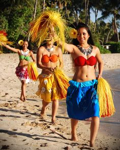 three women in colorful costumes walking on the beach with palm trees and water behind them