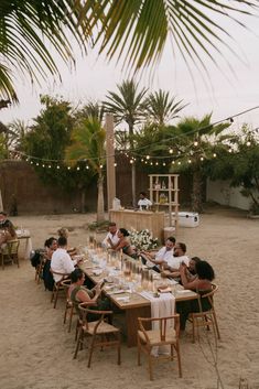 a group of people sitting around a table on top of a sandy beach next to palm trees