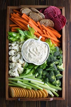 an assortment of vegetables and crackers in a wooden box with dip on the side