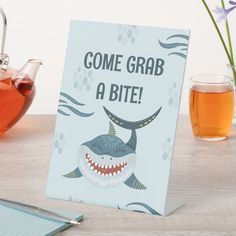 there is a card with a shark on it and some tea in the background that says come grab a bite