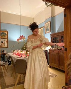 a woman in a white dress standing next to a dining room table with candles on it