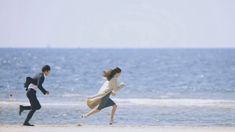 two people are running on the beach near the water and one is wearing a coat