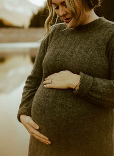 a pregnant woman standing next to a lake with her hands on her stomach and looking down