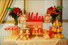 a table topped with lots of cakes and cupcakes next to a window covered in curtains