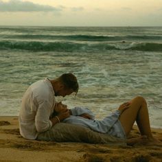 a man and woman are sitting on the sand by the ocean with their heads touching each other