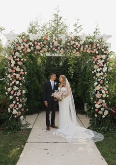 a bride and groom are standing under an arch with flowers on the side, surrounded by greenery