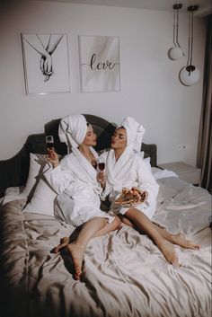 two women in white robes sitting on a bed drinking wine and eating pizza while holding glasses of wine